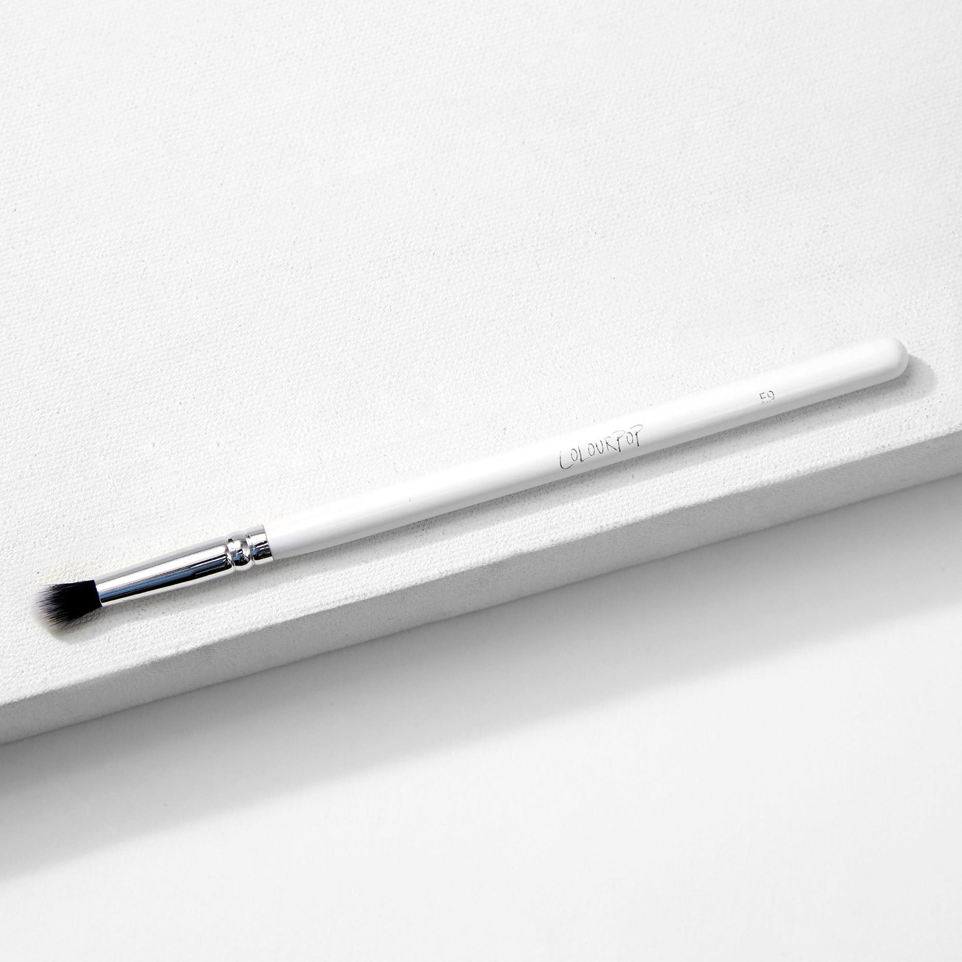ColourPop Small Tapered Brush resting diagonally on a white textured platform, featuring a slim white handle, chrome ferrule, and small tapered black-and-white bristles for precise eyeshadow blending and detail work.