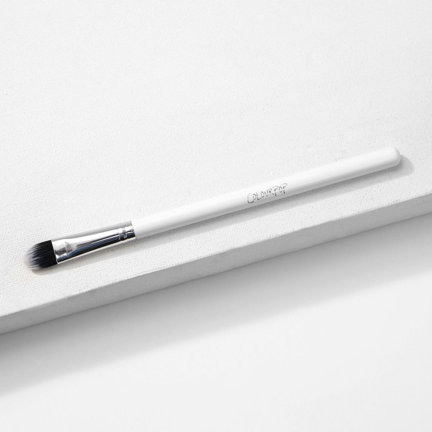 ColourPop Medium Shader Brush with flat oval, dual-toned black-to-white synthetic bristles and a silver ferrule on a white handle, resting on a white textured platform in studio lighting; makeup brush for precise eyeshadow packing and blending.