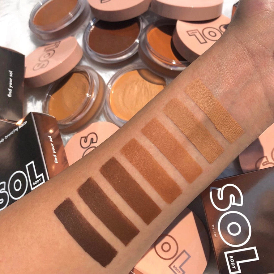 Forearm displays shade swatches of ColourPop SOL Face & Body Bronzing Balm, highlighting the Medium shade among deep to tan tones, against a flat lay of open SOL compacts with blush-pink lids and brown boxes on a white surface.
