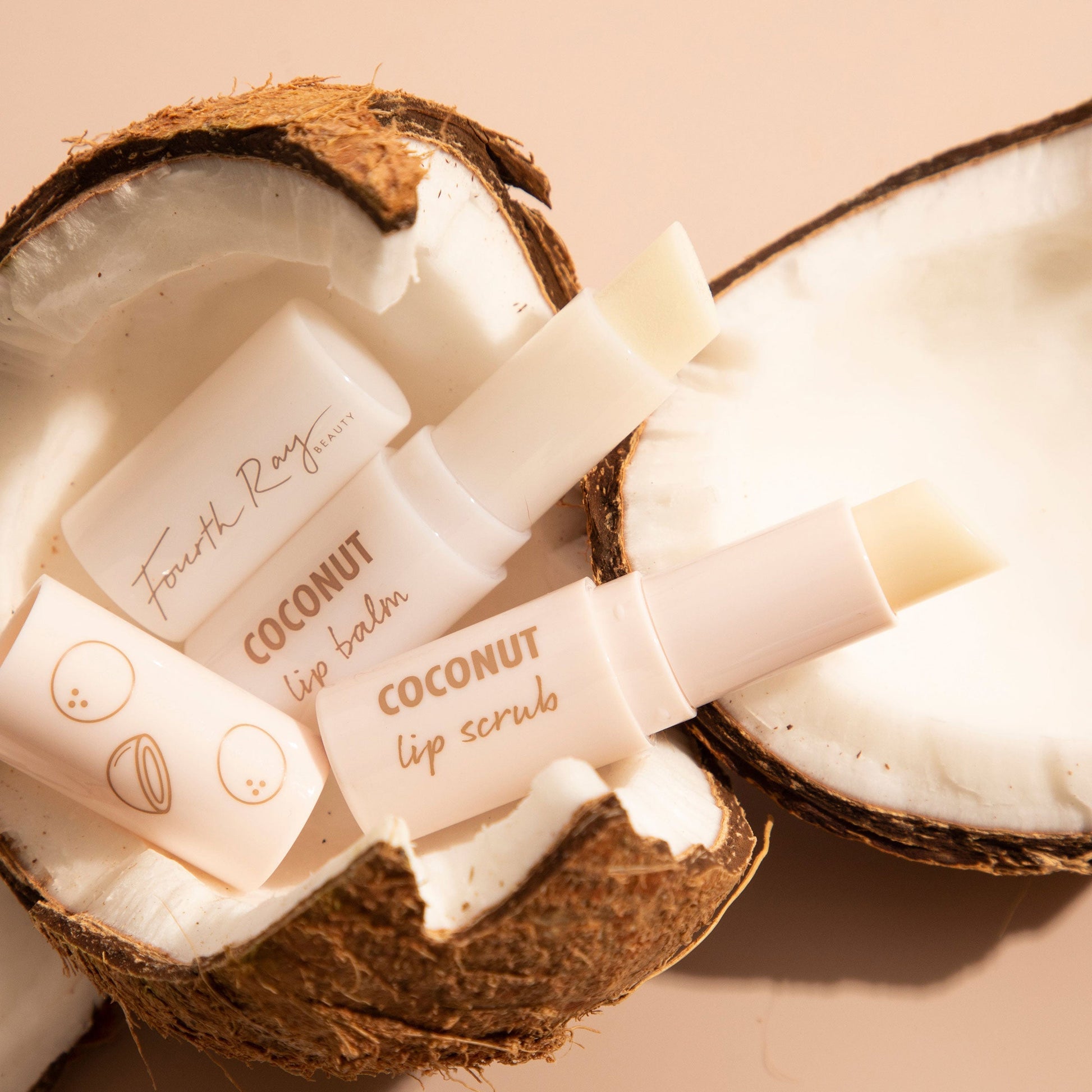 Coco Kisses ColourPop Lip Care Set showing Fourth Ray Beauty coconut lip balm and lip scrub sticks, uncapped and angled, cradled in cracked coconut halves on a soft beige surface, highlighting a tropical, hydrating lip care duo.