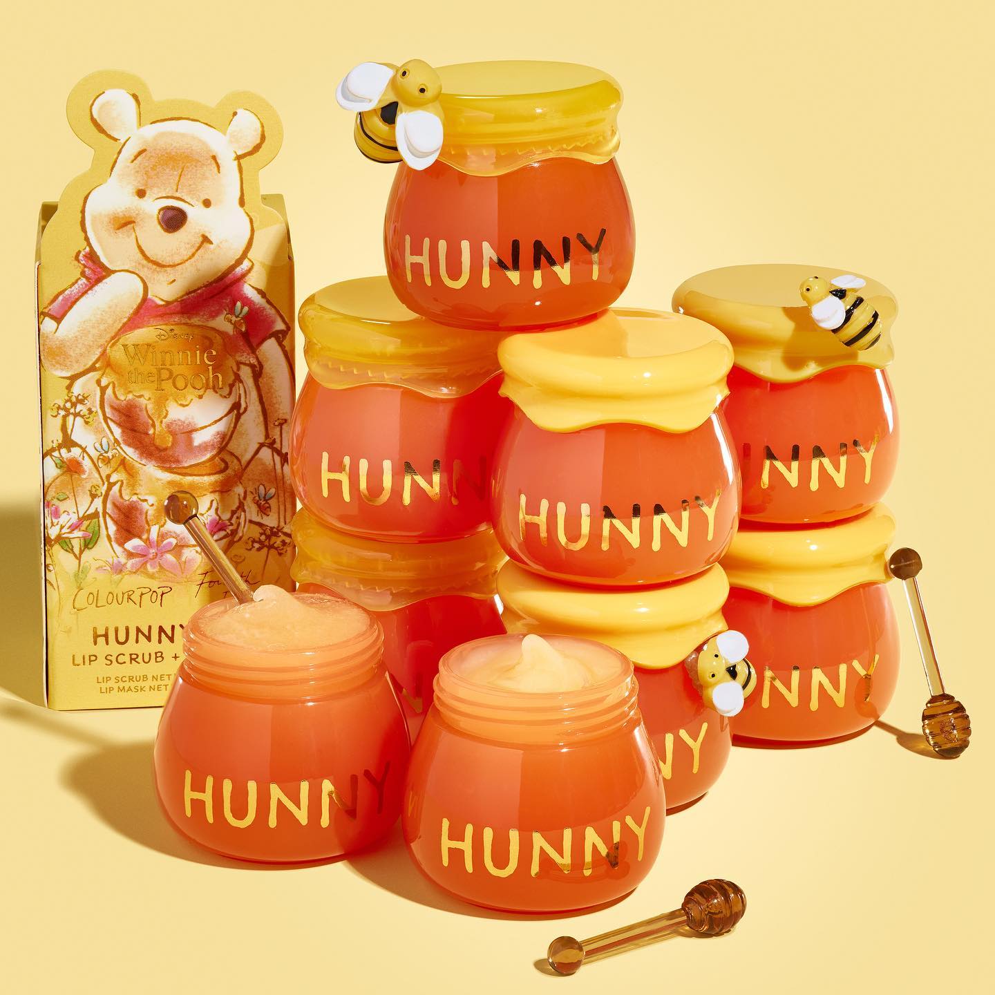 Hunny Pot Lip Care Kit by ColourPop - honey-pot shaped lip care jars stacked and a couple opened to show creamy product, with bee-topped yellow lids, honey dipper applicators, and a Winnie the Pooh box on a pale yellow background.