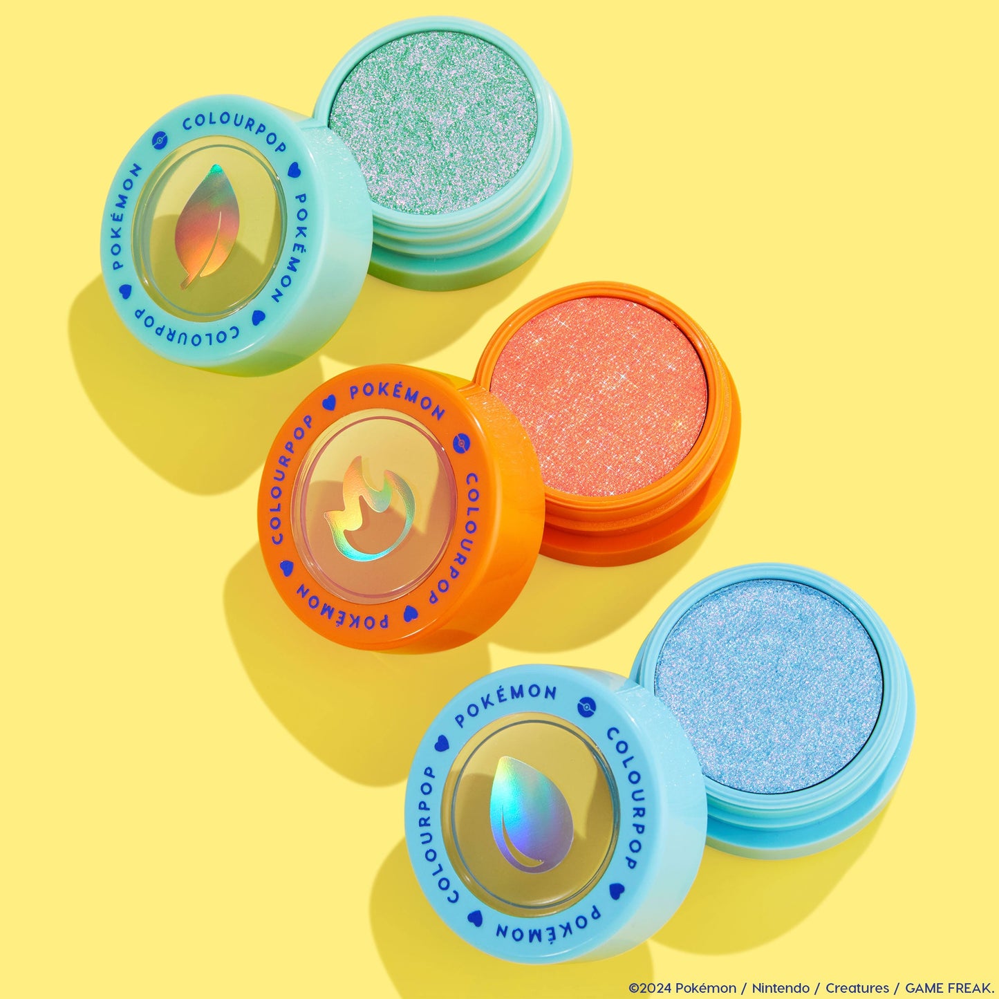 Solar Beam Super Shock Shadow by ColourPop, an open mint-green shimmer pan in an aqua pot, sits beside coral and icy blue companion shades with holographic leaf, flame, and water icons, arranged in a bright yellow flat lay from the Pokémon collection.