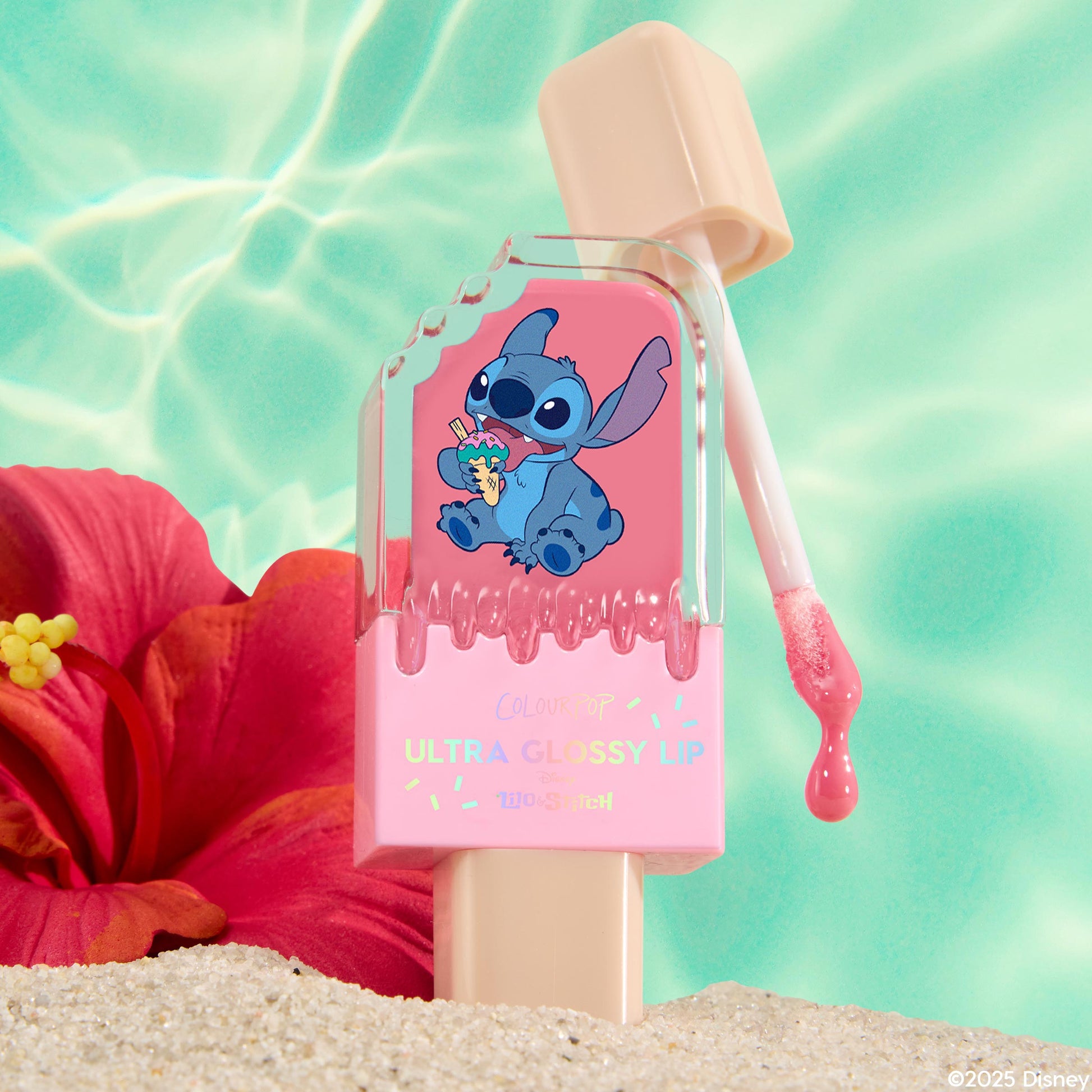 ColourPop Ultra Glossy Lip in Super Sweet stands in beach sand with a doe-foot wand dripping pink gloss; popsicle-style packaging shows Stitch artwork, framed by a turquoise pool backdrop and a red hibiscus for a playful summer vibe.