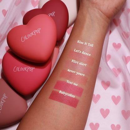 Swatched forearm displays “Text Me” ColourPop Pressed Powder Blush, a rosy pink matte stripe, alongside shades Babycakes, 4ever Yours, Flirt Alert, Let’s Dance, and Kiss N Tell; heart-shaped ColourPop compacts on pink heart-pattern fabric.