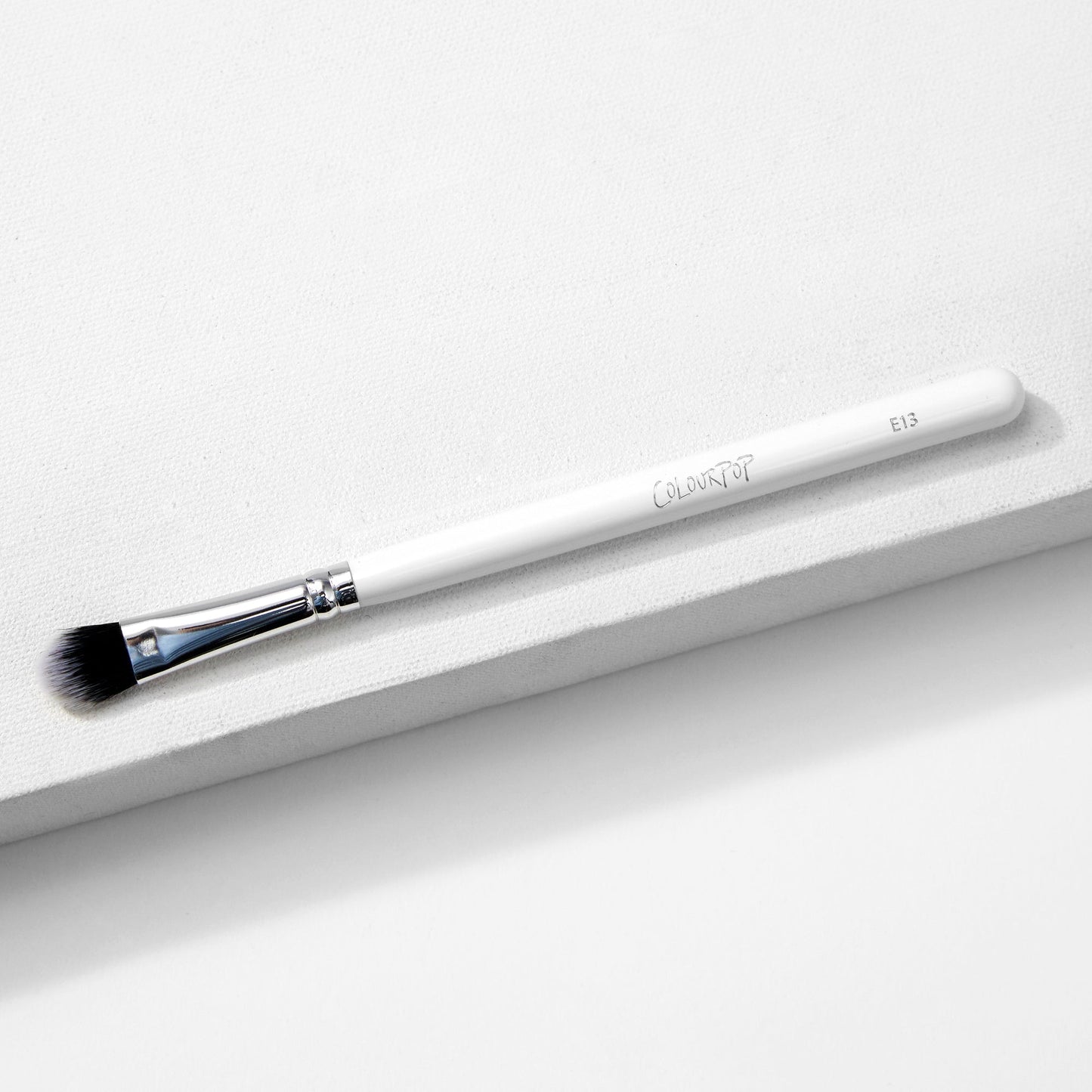 Large Shader Brush makeup brush by ColourPop lies diagonally on a white textured surface, showcasing a white handle with logo and E13, a shiny silver ferrule, and flat dense black-to-white bristles designed for packing and shading eyeshadow.
