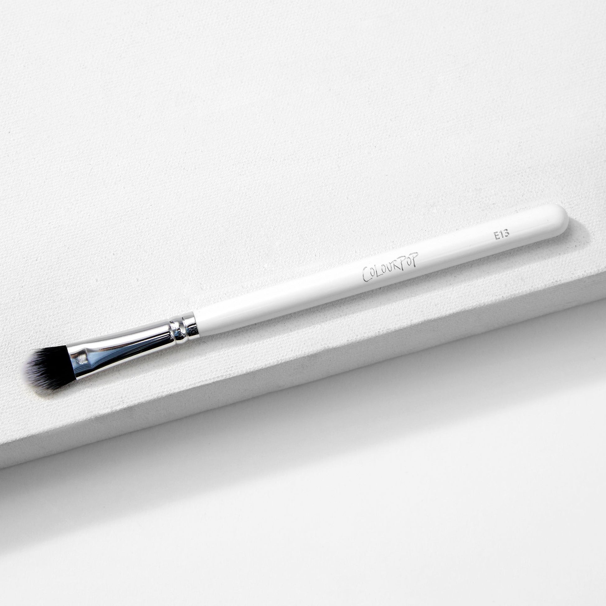 Large Shader Brush makeup brush by ColourPop lies diagonally on a white textured surface, showcasing a white handle with logo and E13, a shiny silver ferrule, and flat dense black-to-white bristles designed for packing and shading eyeshadow.