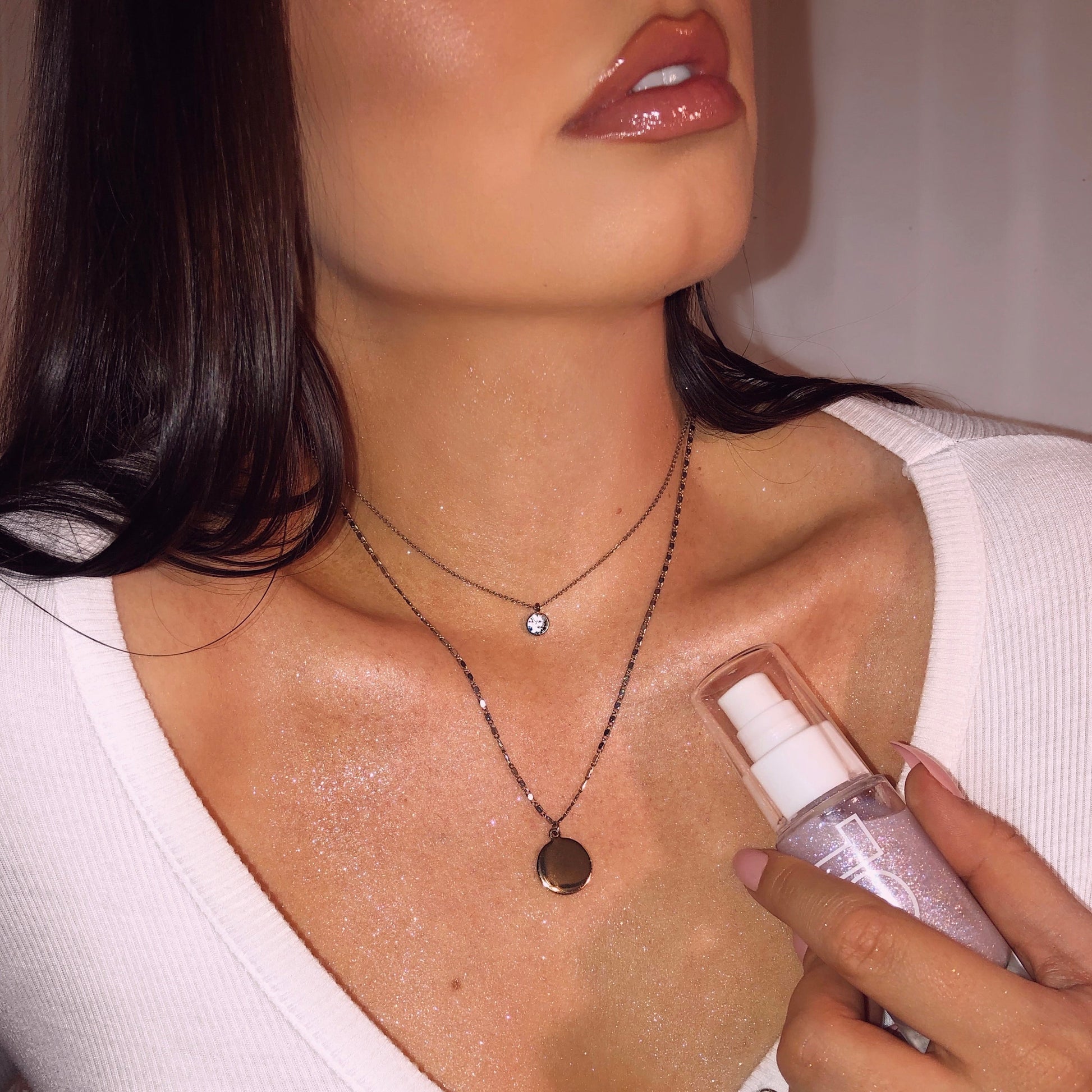 ColourPop SOL Shimmering Dry Oil Opaltini bottle held by a manicured hand as shimmering lavender oil illuminates the model’s collarbones and chest; layered silver necklaces and a white V-neck top frame the radiant glow.